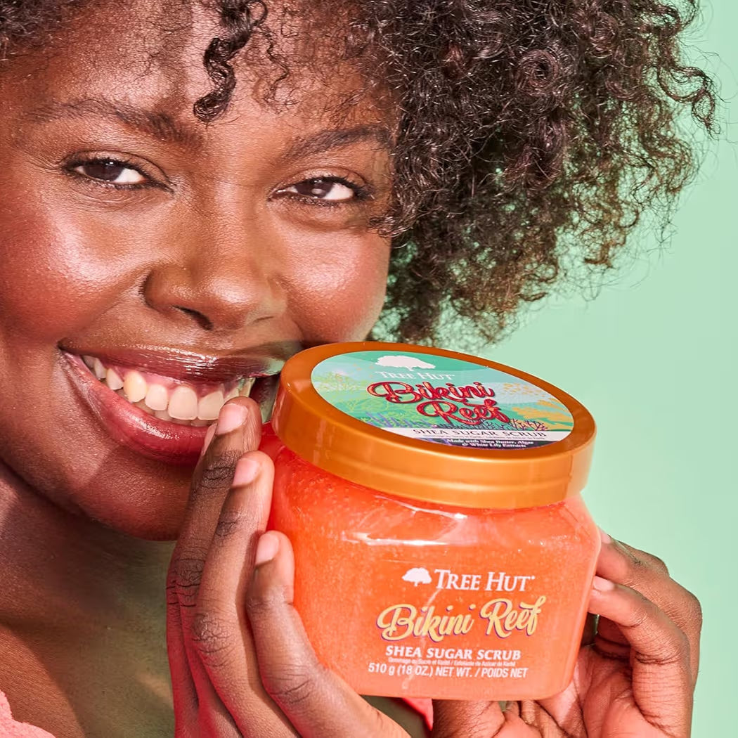HAMYNAT model smiling while holding a jar of Tree Hut Bikini Reef Shea Sugar Scrub against a light green background.