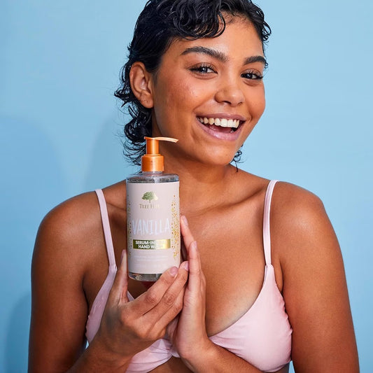 HAMYNAT model smiling while holding a bottle of Tree Hut Vanilla serum-infused hand wash against a blue background